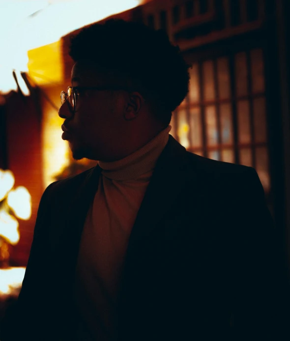 a man in a suit and tie standing in front of a building, an album cover, by Carey Morris, pexels contest winner, renaissance, underexposed lighting, man with glasses, dark skinned, (golden hour)