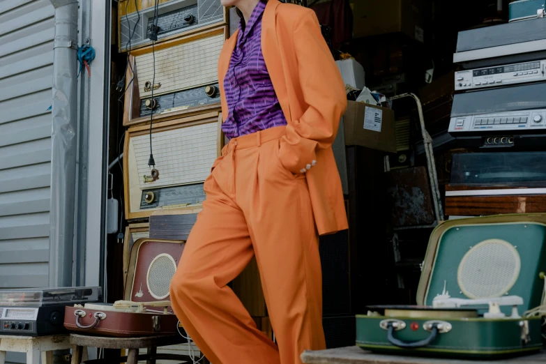 a man in an orange suit standing next to a pile of suitcases, an album cover, by Lee Loughridge, trending on pexels, renaissance, a purple suit jacket, female model, large pants, vintage clothing