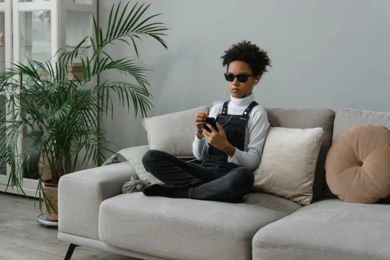 a woman sitting on a couch using a cell phone, by Adam Marczyński, trending on pexels, afrofuturism, television sunglasses, black teenage boy, wearing overalls, with grey skin