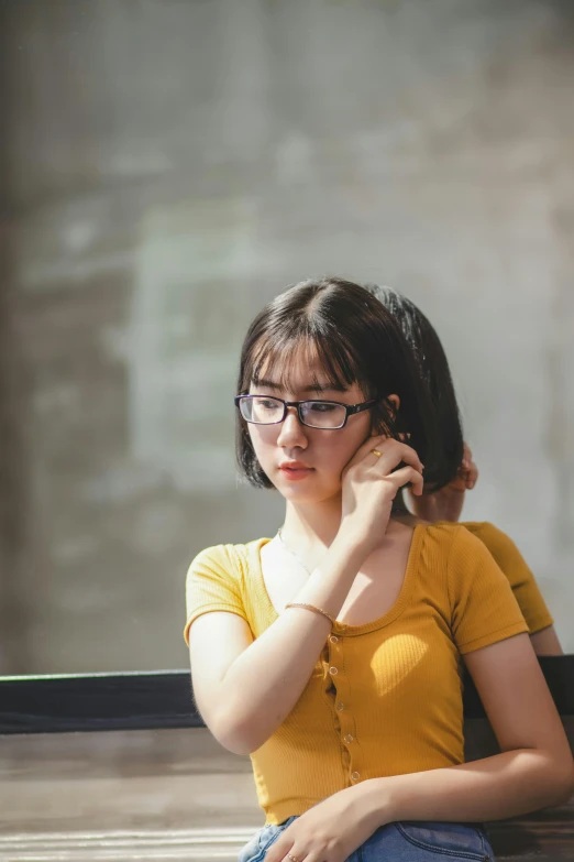 a woman sitting on a bench talking on a cell phone, by Jang Seung-eop, trending on pexels, realism, wearing a modern yellow tshirt, wearing square glasses, serious focussed look, in front of a computer