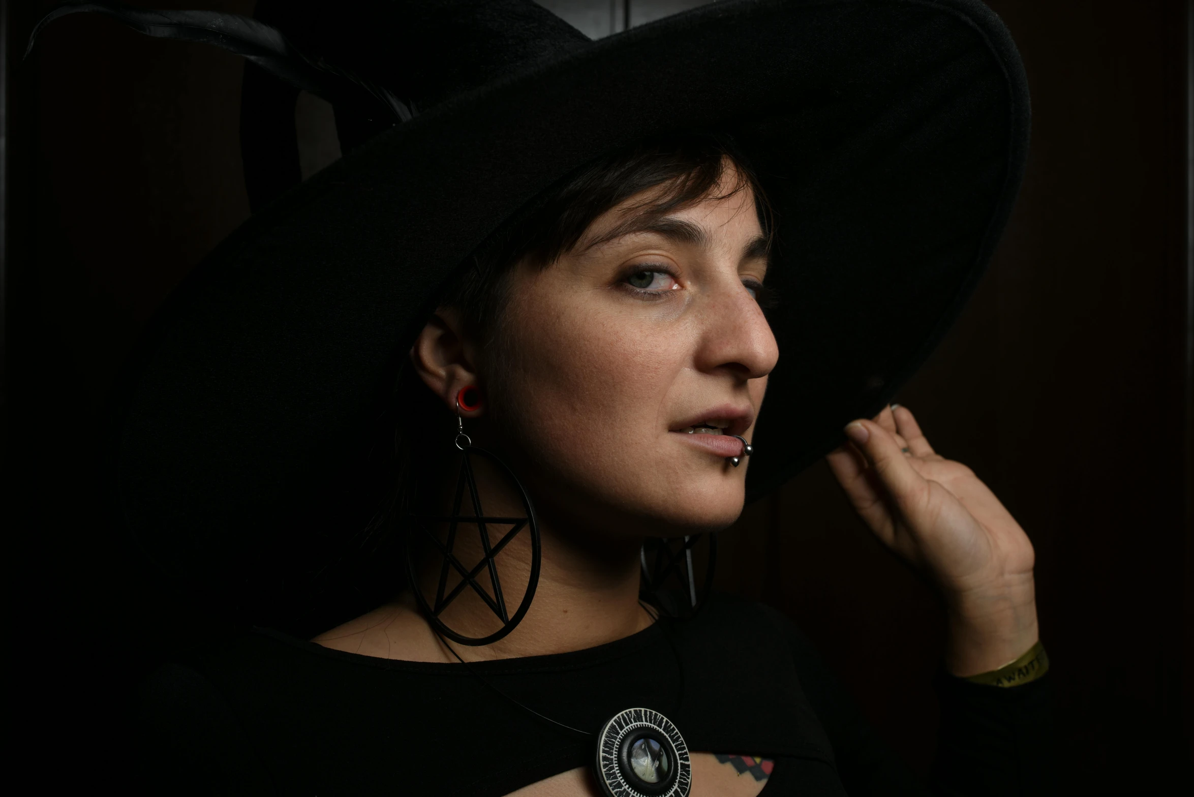 a close up of a person wearing a hat, a character portrait, by Lisa Milroy, pexels contest winner, lowbrow, wearing black witch hat, wearing several pendants, simona sbaffi is the captain, press shot