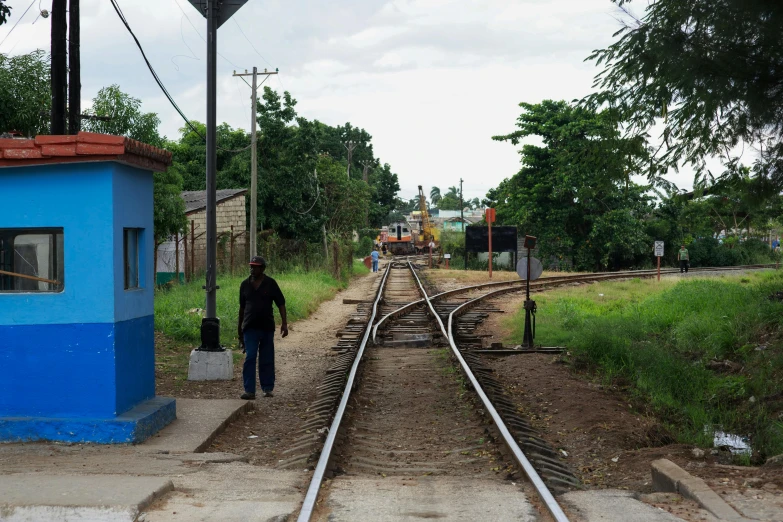 a man walking down a train track next to a blue building, múseca illil, maintenance photo, square, thumbnail