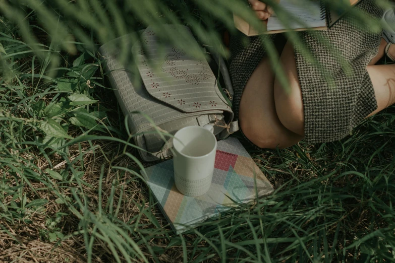 a woman laying in the grass next to a white cup and a coffee mug