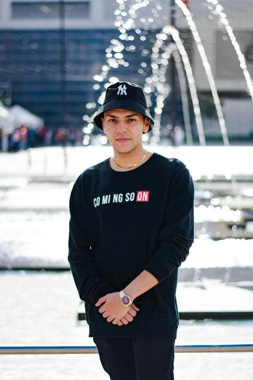 a young man standing in front of a fountain, trending on unsplash, wearing a black sweater, wearing a baseball hat, official store photo, alex yanes