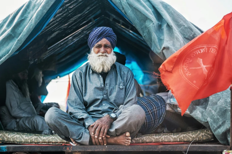 a man sitting on top of a bed in a tent, a portrait, pexels contest winner, on an indian street, japanesse farmer, overalls and a white beard, temporary emergency shelter