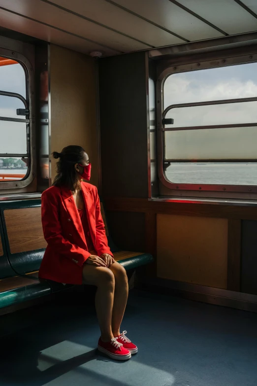 a woman sitting on a bench looking out a window, unsplash contest winner, interior of staten island ferry, red suit, wearing a mask, train far