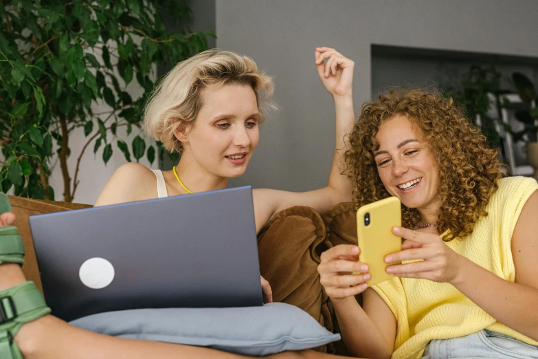 two women sitting on a couch looking at a laptop, trending on pexels, wearing yellow croptop, short curly blonde haired girl, hold up smartphone, avatar image