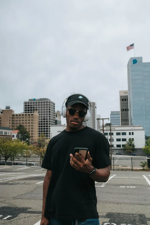 a man standing in a parking lot holding a cell phone, an album cover, by Austin English, unsplash, happening, wearing sunglasses and a cap, city view, alabama, dark skinned