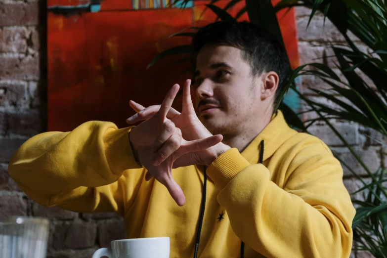 a man sitting at a table with a cup of coffee, wearing a yellow hoodie, hand gesture, liam brazier, avatar image