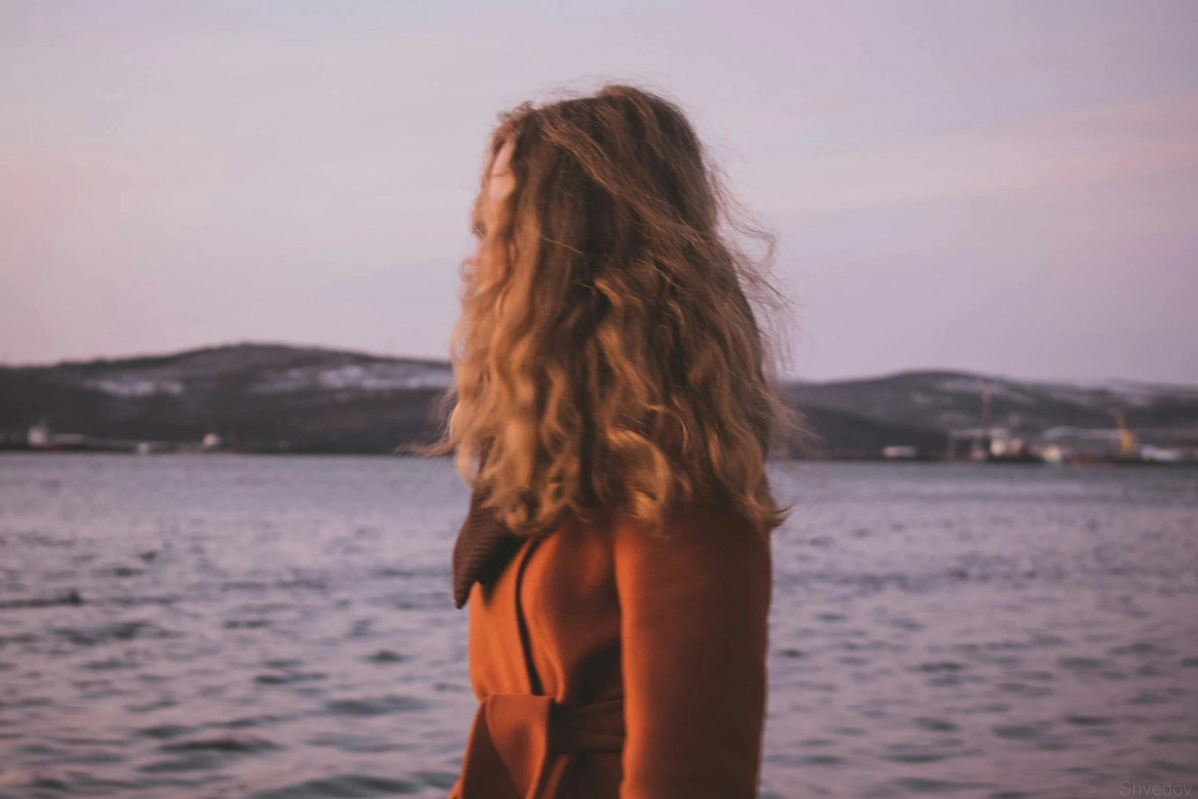 a woman standing in front of a body of water, a picture, inspired by Elsa Bleda, trending on pexels, renaissance, curly dirty blonde hair, faded red colors, looking into the distance, purple hue