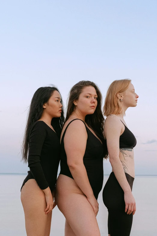 a group of women standing next to each other on a beach, an album cover, inspired by Vanessa Beecroft, unsplash, renaissance, full - bodied portrait, profile image, three women, wearing a black bodysuit