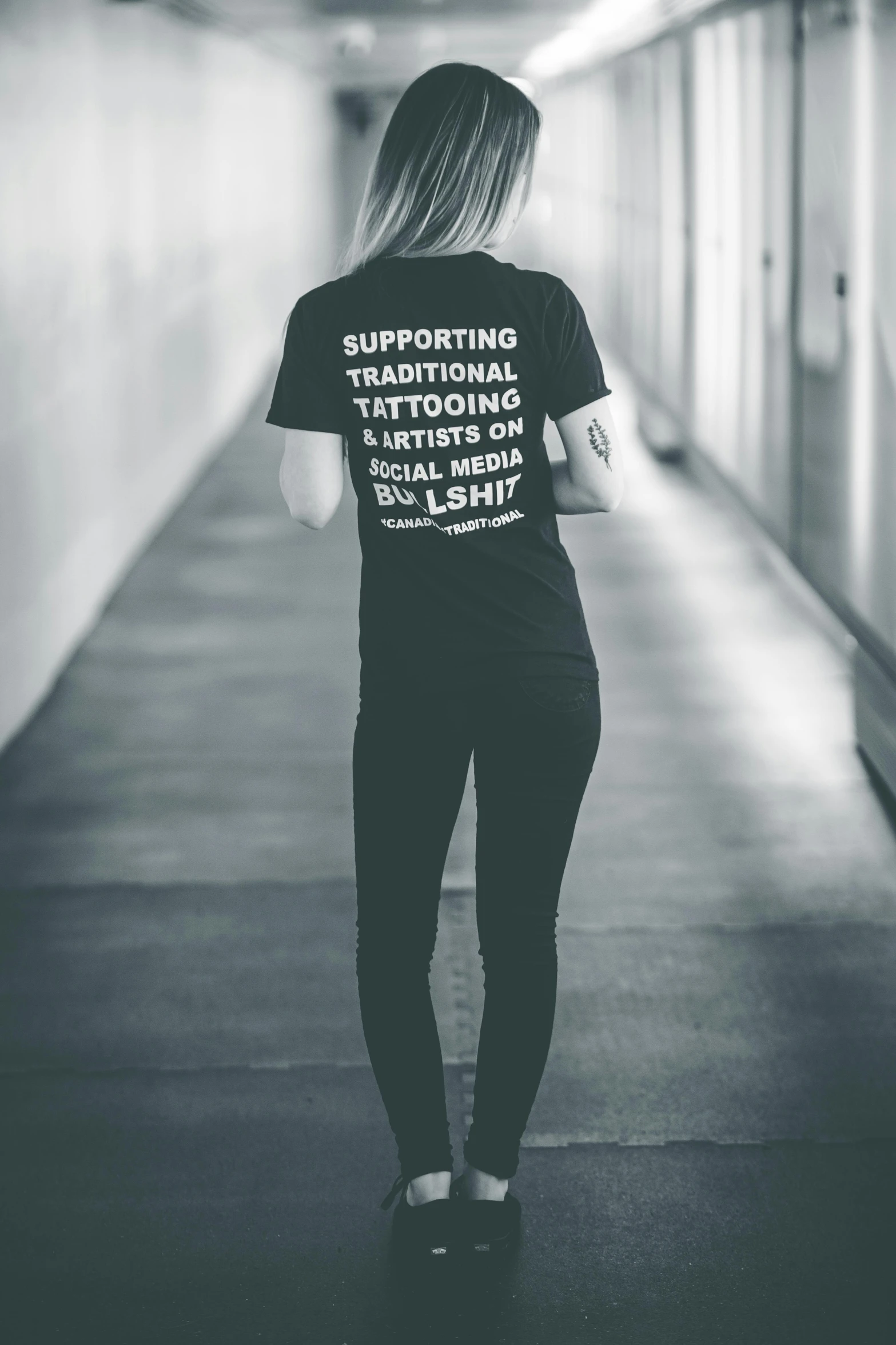 a woman in a black shirt is walking down a hallway, a tattoo, unsplash contest winner, t-shirt, support, fullbody photo, on grey background