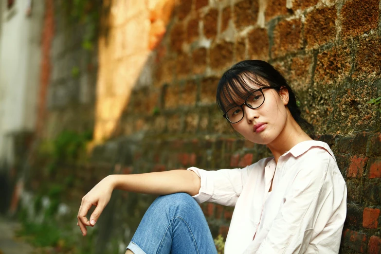 a woman sitting in front of a brick wall, inspired by Pan Yuliang, pexels contest winner, realism, square rimmed glasses, teen girl, asian, thoughtful )