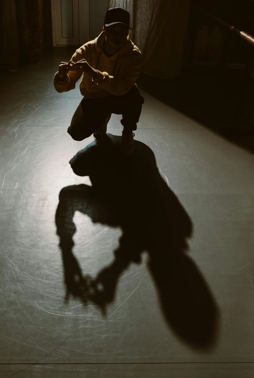 a man kneeling on a skateboard in a dark room, by Paul Bird, cast shadows, capoeira, sun puddle, on an empty stage from above