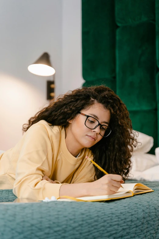 a woman laying on a bed with a notebook and pen, trending on pexels, academic art, yellow clothes, curls, nerds, light skin