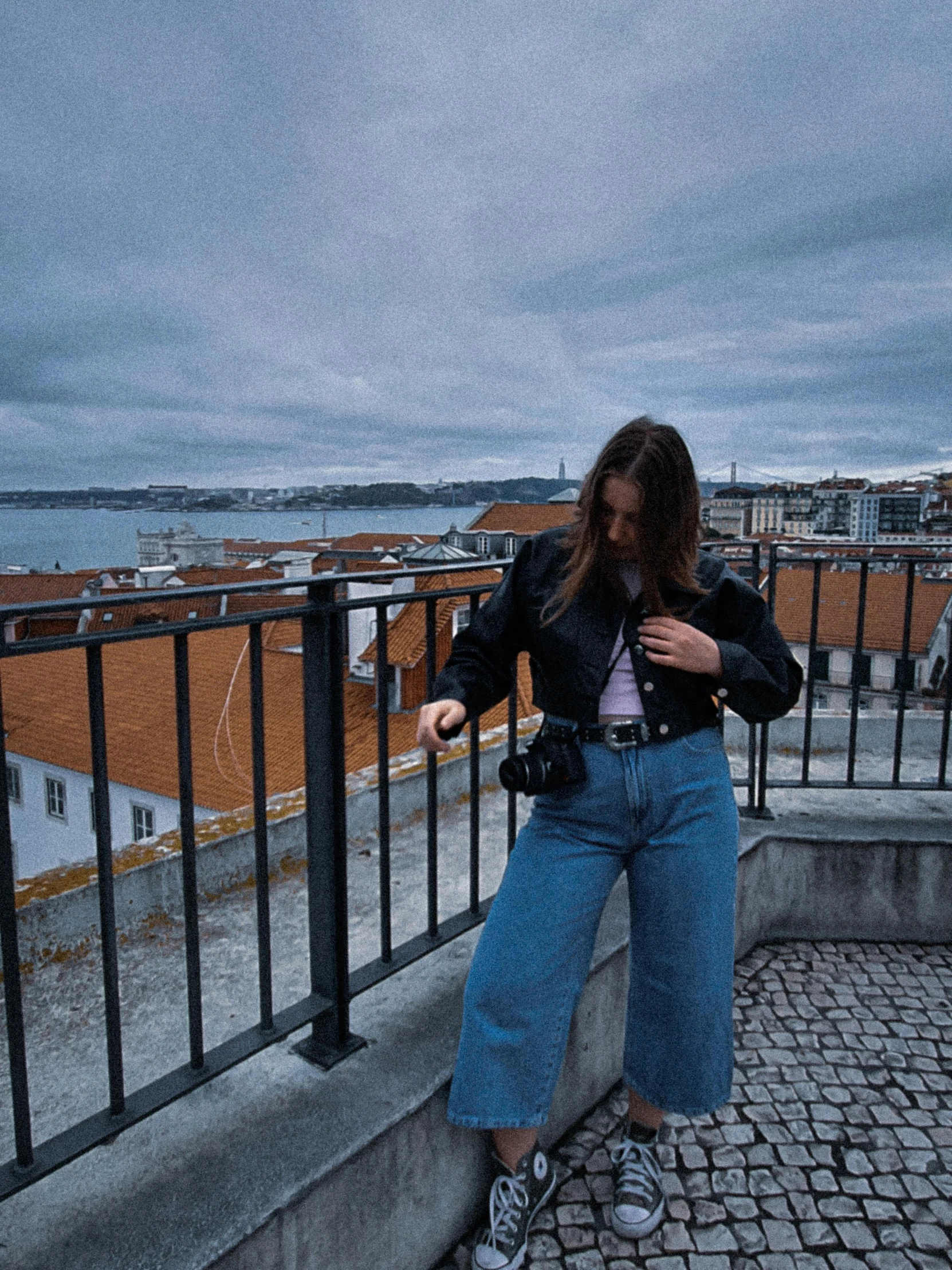 a woman standing on a ledge looking at her cell phone, by Olivia Peguero, pexels contest winner, lisbon, an aviator jacket and jorts, looking back at the camera, holding a camera