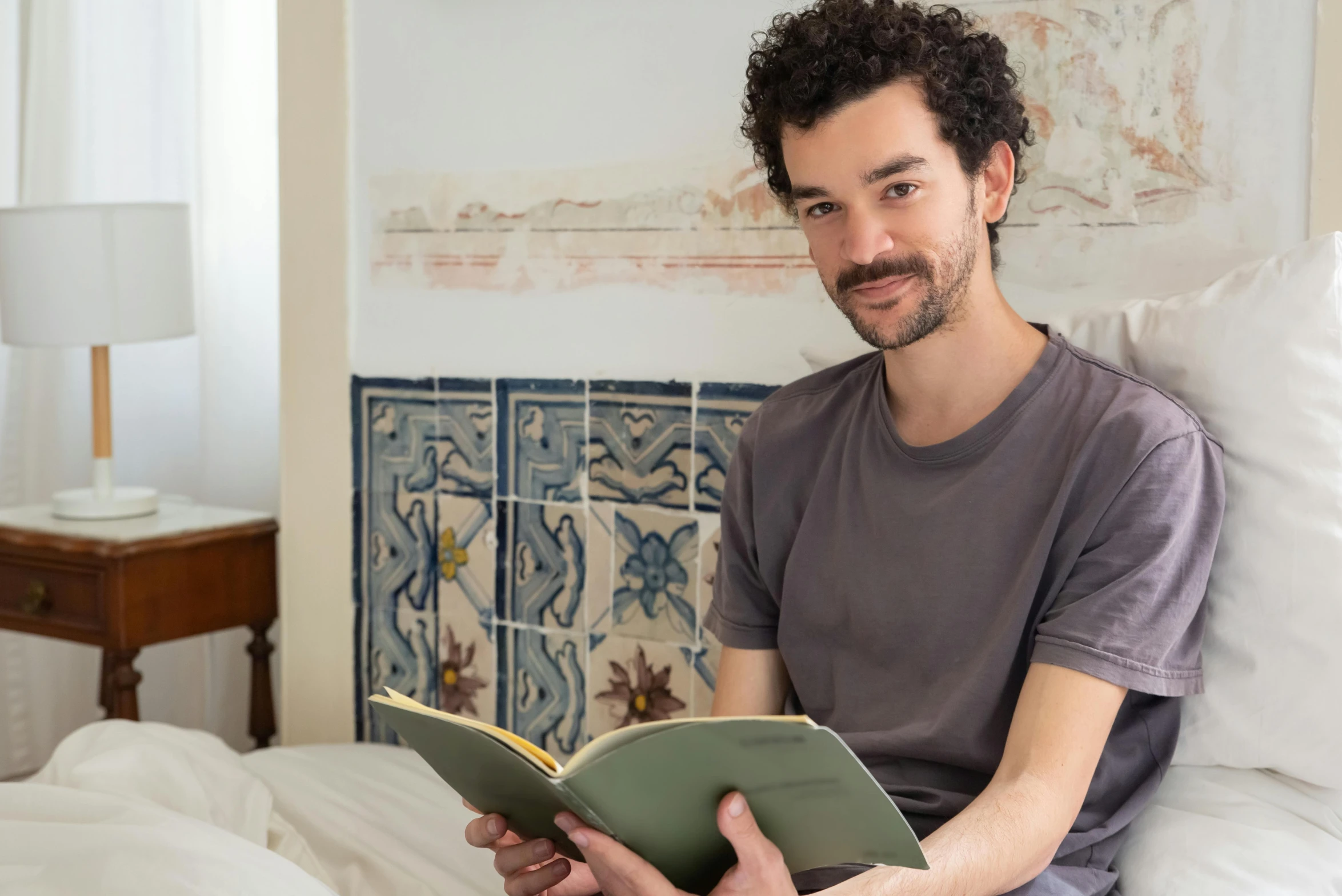 a man sitting on a bed reading a book, inspired by Santiago Rusiñol, happening, looking towards camera, profile image, portrait image, robert sheehan