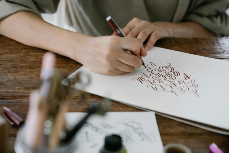 a woman sitting at a table writing on a piece of paper, a drawing, inspired by Art & Language, trending on pexels, “modern calligraphy art, brown, silver and crimson ink, detailed letters