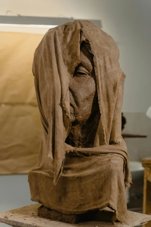 a sculpture of a woman sitting on top of a table, inspired by Käthe Kollwitz, reddit, face covered in dirt, hood covers his eyes, leonardo davinci detail, long flowing fabric