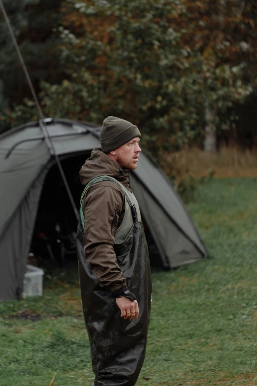 a man standing in a field next to a tent, wearing a fisher 🧥, model is wearing techtical vest, jörmungandr, gloomy mood