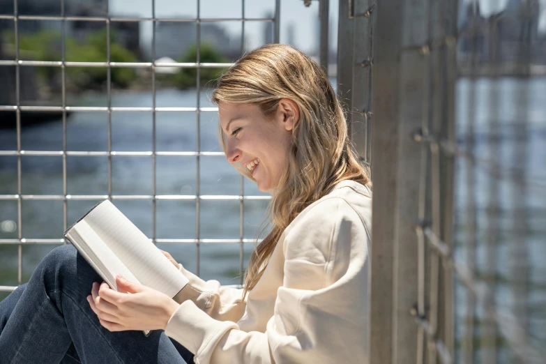 a woman sitting on the ground reading a book, sydney sweeney, on a bridge, smiling laughing, profile image