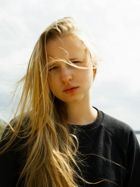 a woman with long blonde hair standing in front of a body of water, an album cover, unsplash, realism, beautiful taissa farmiga, grimacing, greta thunberg, non-binary