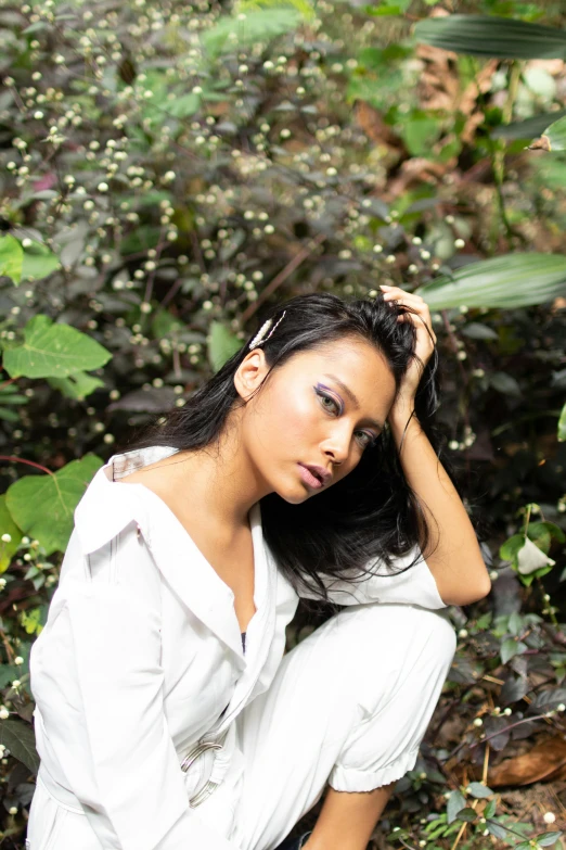 a woman sitting on the ground in the woods, an album cover, by Basuki Abdullah, sumatraism, wearing a white button up shirt, pouting, actress, next to a plant