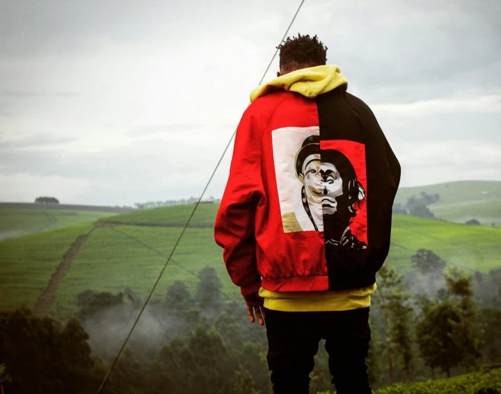 a man standing on top of a lush green hillside, an album cover, by Afewerk Tekle, trending on pexels, visual art, red hoodie, wiz khalifa, black and yellow and red scheme, bomber jacket