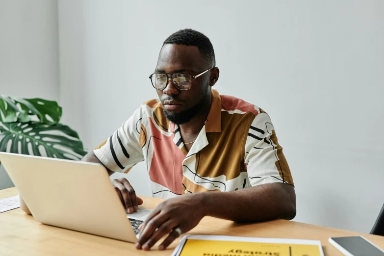 a man sitting at a table using a laptop computer, by Carey Morris, trending on pexels, afrofuturism, wearing round glasses, brown, thumbnail, professional photo