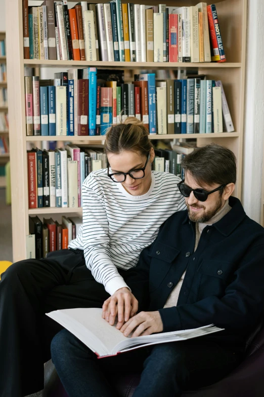 a man and a woman sitting on a couch in front of a bookshelf, by Nina Hamnett, trending on unsplash, academic art, with nerdy glasses and goatee, academic clothing, russian academic, sat at a desk