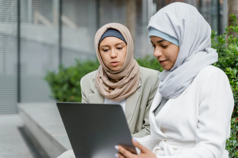 two women sitting on a bench looking at a laptop, a picture, trending on pexels, hurufiyya, islamic, pictured from the shoulders up, holding notebook, thumbnail