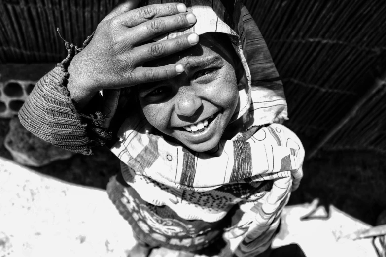 a black and white photo of a smiling child, by Ahmed Yacoubi, pexels contest winner, blinding bright sun, covered head, 🤤 girl portrait, greeting hand on head