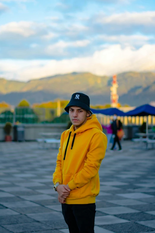 a man in a yellow jacket is standing on a skateboard, unsplash, hat and hoodie, standing in front of a mountain, russian and japanese mix, david marquez