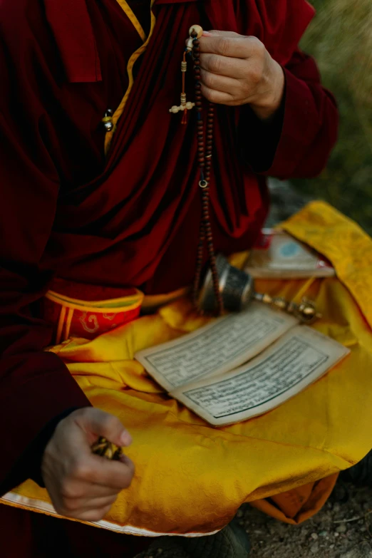 a man that is sitting down with a rosary, pexels contest winner, hurufiyya, tibetan text script, wearing red and yellow clothes, holding a book, wearing dark robe