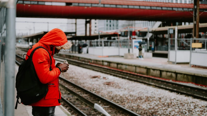 a woman standing on a train platform looking at her phone, by Andries Stock, pexels contest winner, graffiti, orange hoodie, red cloth around his shoulders, wearing a red gilet, outside alone smoking weed