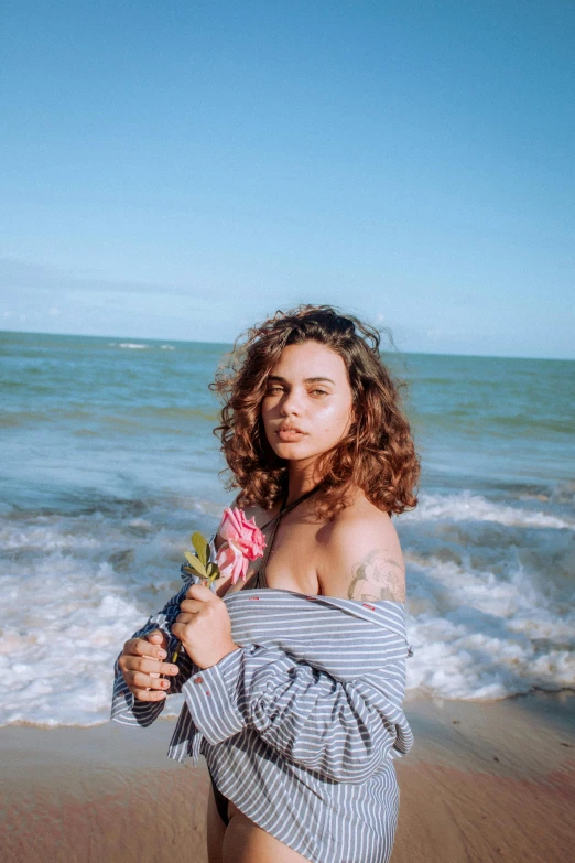 a woman standing on top of a beach next to the ocean, brown curly hair, photo of a rose, androgynous person, caio santos