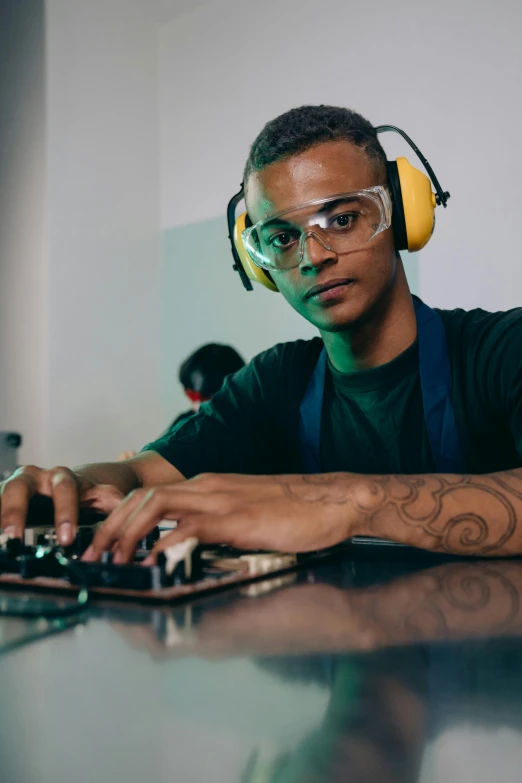 a man sitting at a table with headphones on, inspired by Afewerk Tekle, trending on pexels, arbeitsrat für kunst, portrait of computer & circuits, wearing overalls, wearing goggles, teenage engineering moad