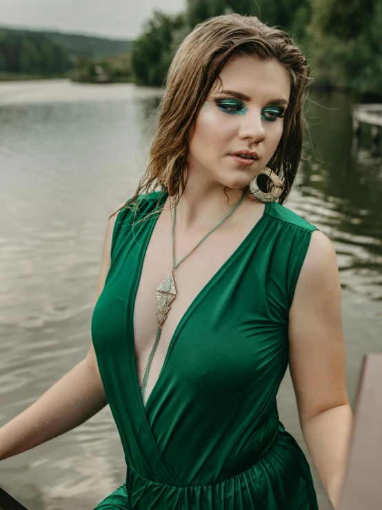 a woman in a green dress standing next to a body of water, an album cover, inspired by Elsa Bleda, unsplash, renaissance, neck chains, cleavage, high resolution product photo, model posing