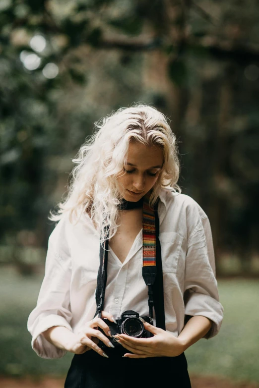 a woman in a white shirt and black pants holding a camera, by Tom Bonson, unsplash contest winner, curly blonde hair, lush surroundings, sydney sweeney, white hair dreads