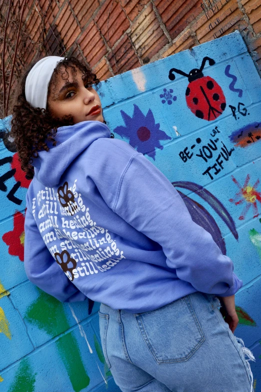 a woman standing in front of a wall covered in graffiti, an album cover, graffiti, blue hoodie, ((blue)), pastel clothing, profile image