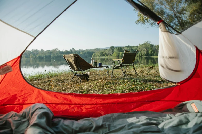 a red and white tent sitting next to a body of water, by Jessie Algie, trending on unsplash, two plastic chair behind a table, sleeping bag, sydney park, interior of a tent