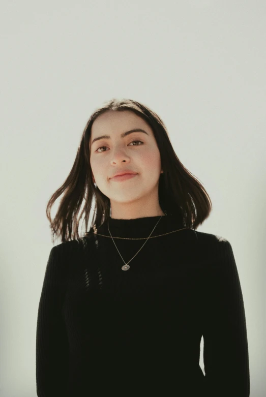 a woman standing in front of a white wall, wearing a black sweater, charli bowater, straight eyebrows, arm around her neck