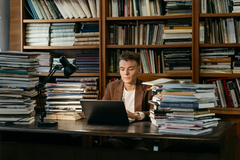 a woman sitting at a desk in front of a laptop computer, by Julia Pishtar, pexels, academic art, shelves filled with tomes, joe keery, lachlan bailey, julian ope