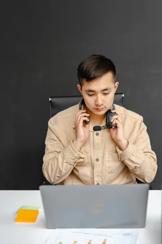 a man sitting at a table with a laptop and a cell phone, wearing a headset, dao trong le, high-quality photo, thumbnail