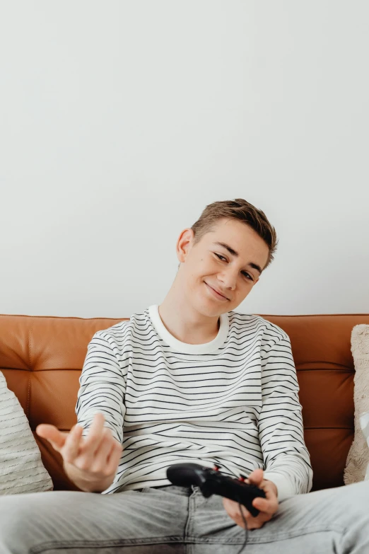 a man sitting on a couch holding a video game controller, an album cover, by Carey Morris, trending on pexels, happening, portrait androgynous girl, wearing stripe shirt, julian ope, laughing