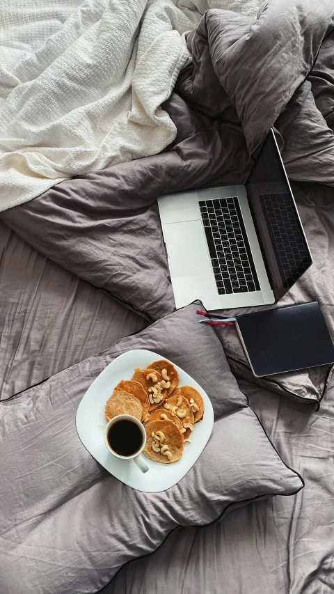 a bed with a plate of food, laptop and mug on it