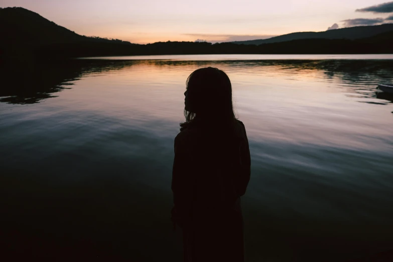 a person standing in front of a body of water, inspired by Elsa Bleda, pexels contest winner, romanticism, black silhouette, teenage girl, unkept hair, lake in the distance