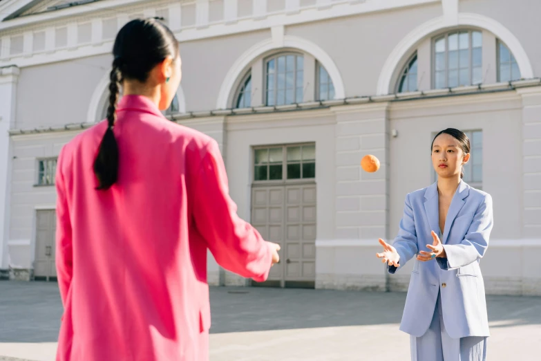 a woman throwing an orange ball to another woman, by Julia Pishtar, pexels contest winner, wearing a light - pink suit, wearing a blue jacket, asian female, official vogue editorial