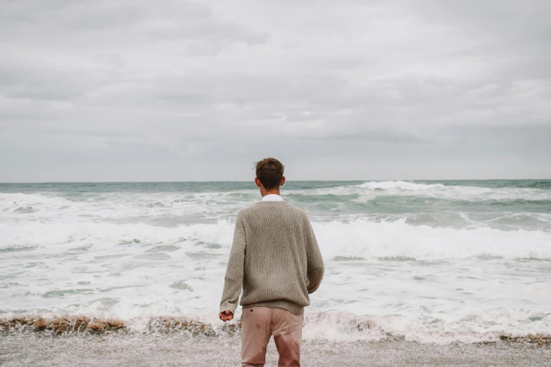 a man standing on top of a beach next to the ocean, wearing sweater, big overcast, background image, ignant