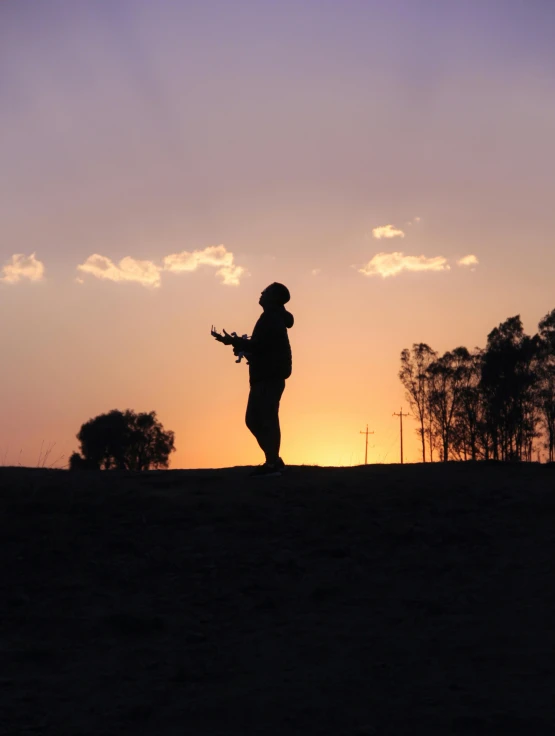 a silhouette of a person standing on top of a hill, an album cover, pexels contest winner, holding a mp7, in the australian outback, standing in the grass at sunset, profile pic